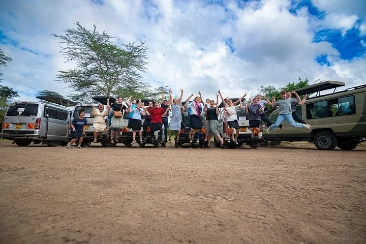 A group of travellers jumping for a photo beside their safari vans on a dirt clearing